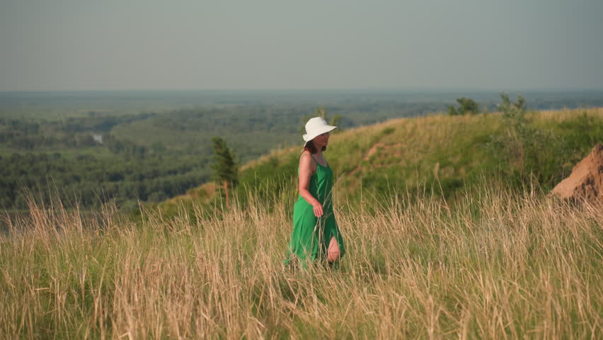 woman in white hat and green gown walks along grassy cliffside, turning to gaze across lush valley landscape with forest stretching into distant horizon under soft sunlight of clear sky