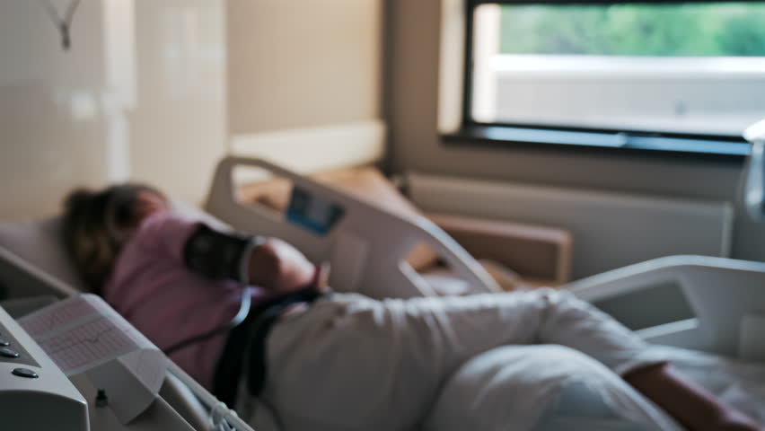 Close-up of a fetal monitor in a doctor's office, displaying fetal heart rate and uterine contraction data during a prenatal check-up with a blurred view of a pregnant woman lying in a hospital bed - Powered by Shutterstock - Get 15% off with code: PIKWIZARD15