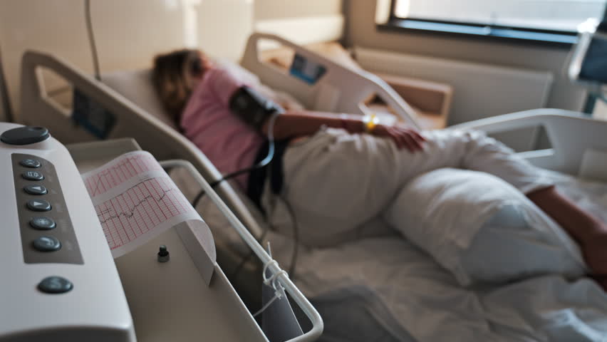 Close-up of a fetal monitor in a doctor's office, displaying fetal heart rate and uterine contraction data during a prenatal check-up with a blurred view of a pregnant woman lying in a hospital bed - Powered by Shutterstock - Get 15% off with code: PIKWIZARD15