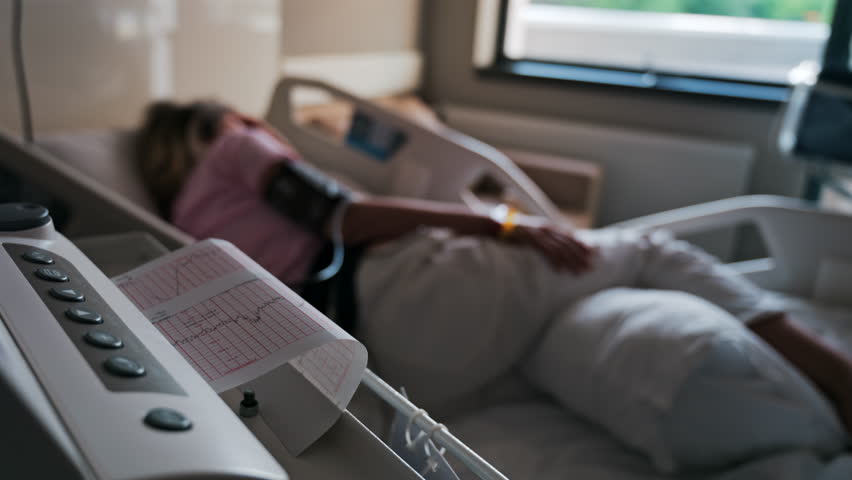 Close-up of a fetal monitor in a doctor's office, displaying fetal heart rate and uterine contraction data during a prenatal check-up with a blurred view of a pregnant woman lying in a hospital bed - Powered by Shutterstock - Get 15% off with code: PIKWIZARD15