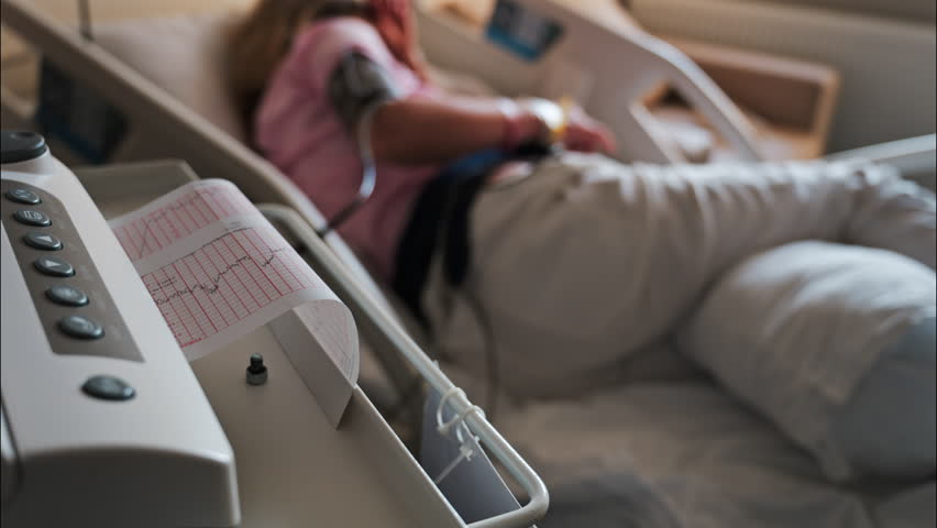 Close-up of a fetal monitor in a doctor's office, displaying fetal heart rate and uterine contraction data during a prenatal check-up with a blurred view of a pregnant woman lying in a hospital bed - Powered by Shutterstock - Get 15% off with code: PIKWIZARD15