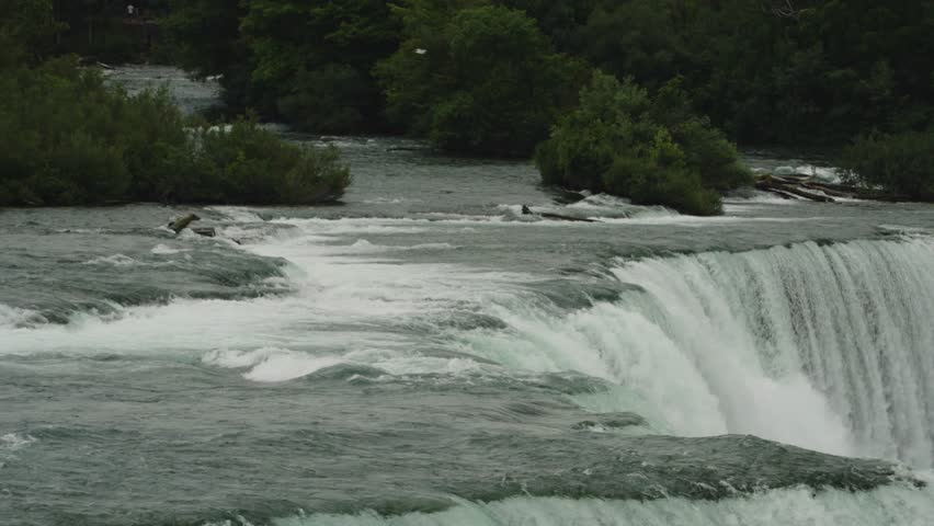 Close-up of Niagara River rushing over the edge of the falls with lush greenery framing the scene and turbulent water cascading into the gorge.