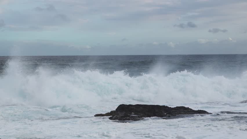 A heavy shorebreak slams into a dark rock and detonates into white spray while churning foam floods the foreground beneath pale clouds and a steady onshore breeze.