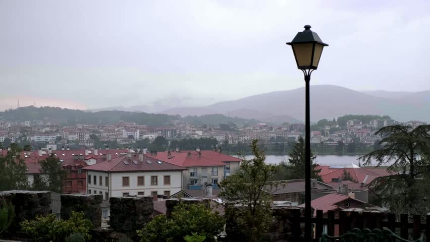 Overcast view of Hondarribia rooftops and streetlamp with misty hills across the river in France.