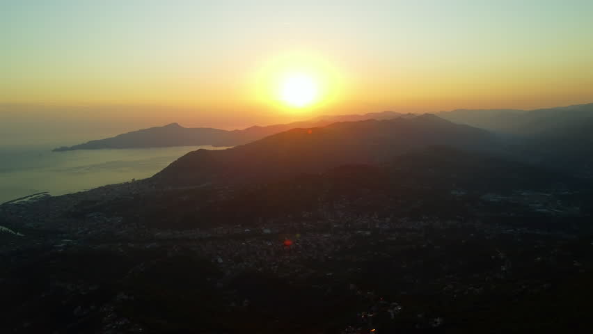 4K aerial drone footage flying forward at sunset, capturing a red-orange sun beyond forested mountains above a city below, for cinematic and dramatic landscape.