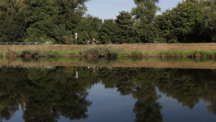 Cyclists riding along a riverside path with lush green trees reflecting in calm water under clear summer sky creating peaceful outdoor recreation and family leisure atmosphere