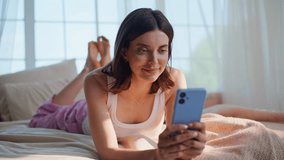 Girl scrolling news feed at cellphone lying comfortable home bed closeup. Relaxed brunette messaging smartphone relaxing in bedroom. Carefree woman enjoy online phone communication at domestic weekend - Powered by Shutterstock - Get 15% off with code: PIKWIZARD15