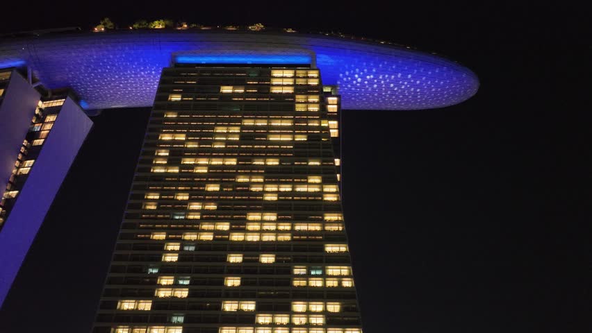Aerial view of Marina Bay Sands shimmers with illuminated windows and a blue-lit rooftop against the night sky, Singapore, Singapore.