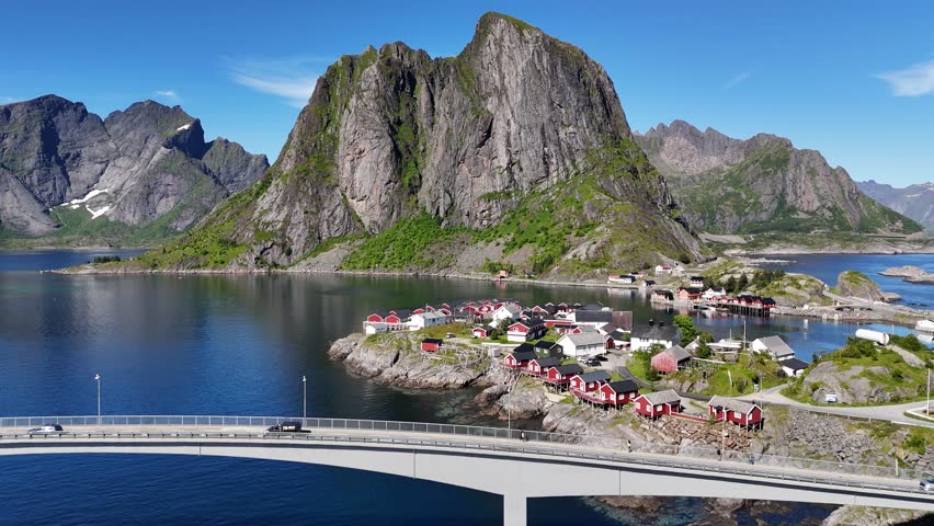 Drone flying over the car bridge and small fishing village Hamnøy in Norway towards iconic mountains above the sea with wooden drying racks for torrfisk