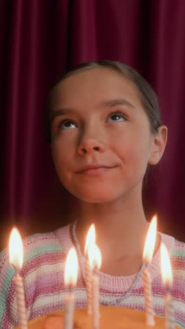 Vertical portrait of cheerful little Asian birthday girl blowing candles on birthday cake and smiling at camera, posing against red curtain background