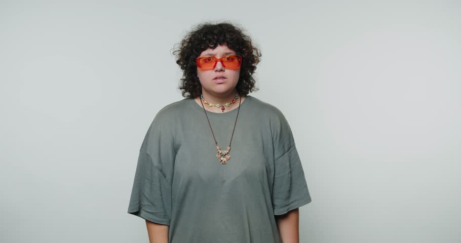 Person with curly hair wearing orange sunglasses making a shushing gesture.