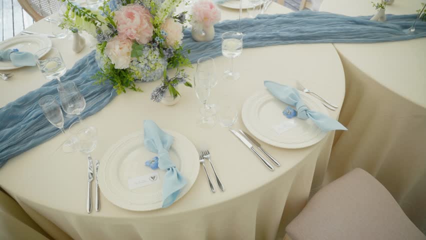 Elegant wedding table set with blue napkins, pastel floral centerpiece, and fine glassware on ivory linen—perfect for a romantic and upscale reception setting.