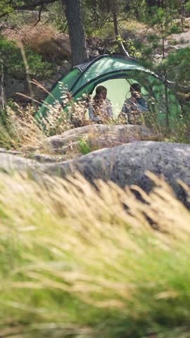 Couple in a green tent among rocks and pine trees in Finland, enjoying peaceful camping in the forest with tall grass in the foreground.