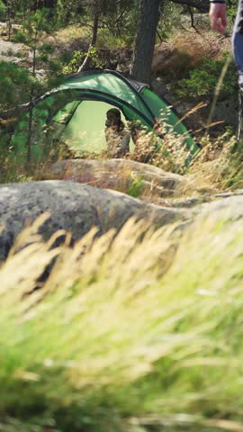 Couple in a green tent among rocks and pine trees in Finland, enjoying peaceful camping in the forest with tall grass in the foreground.