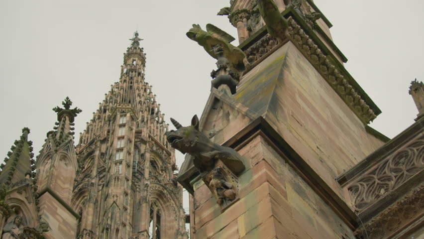 Close-up of Strasbourg Cathedral’s ornate Gothic spire, detailed gargoyles, and stone statues, showcasing medieval craftsmanship against a pale sky.