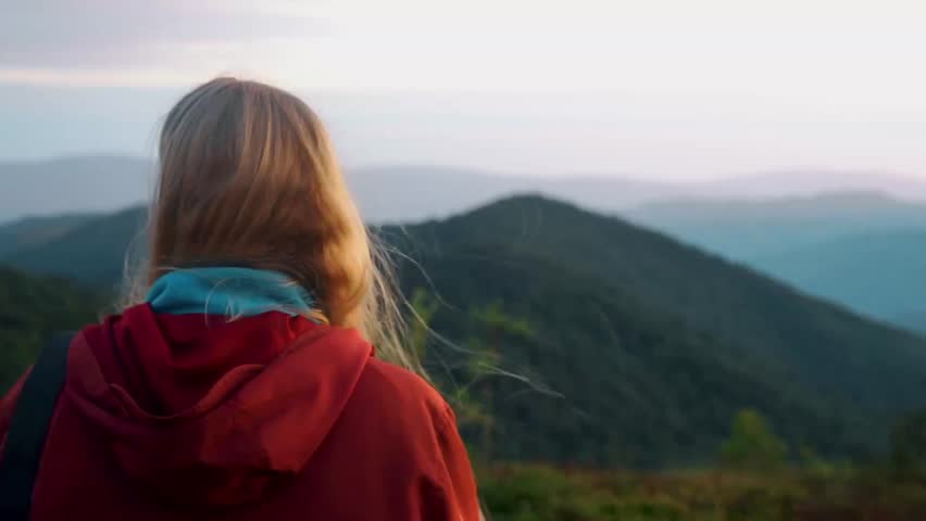 Woman watching the sunset, seen from behind, against a backdrop of mountains at sunset, natural phenomena, in soft warm light, mountain peaks and silhouettes of ridges, travel