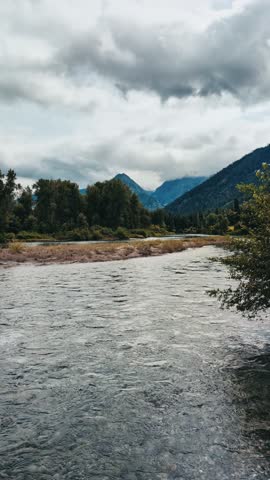 A Vertical shot of a shallow river flowing through a valley with mountains in the background under a cloudy sky