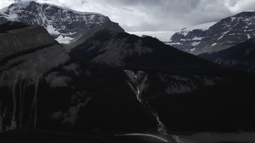 A drone view of a dramatic mountain range with dark, forested slopes and distant snow-capped peaks under a cloudy sky in Alberta, Canada