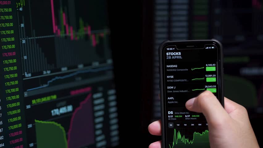 A person uses their smartphone to check stock market trends and financial data, with a computer screen displaying charts and graphs in the background. - Powered by Shutterstock - Get 15% off with code: PIKWIZARD15