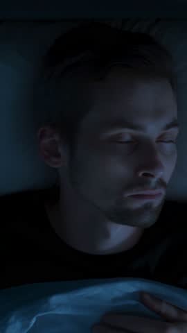 Close-up, in a dark bedroom at night, a man wakes up because of sleep paralysis or nightmare, drained by insomnia and a sleep disorder, overwhelmed by anxiety, fatigue. vertical video