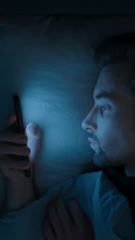 A tired man in bed scrolls endlessly on his mobile phone, struggling with insomnia, anxiety and overthinking as he watches social media late into the night. Smartphone dependence. vertical video