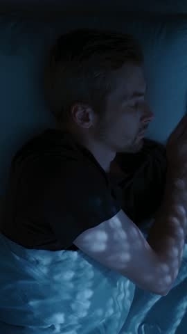 A young man lies awake in bed at night, turning restlessly due to insomnia, frustrated and exhausted, struggling with overthinking and sleep issues. Top view, time lapse. vertical video