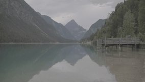 Peaceful view of an Austrian alpine lake with calm turquoise water, wooden pier, and surrounding mountains reflecting on the surface under cloudy sky. Perfect serene nature scenery - Powered by Shutterstock - Get 15% off with code: PIKWIZARD15