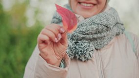 close up of cheerful woman in cozy jacket and thick scarf smiling joyfully while holding bright red autumn leaf between fingers, showcasing beauty of nature during crisp fall - Powered by Shutterstock - Get 15% off with code: PIKWIZARD15