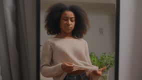 African American woman looking in mirror reflection at home trying beige jumper dissatisfied girl female wearing casual style bad choice going away frustrated with clothes quality terrible outfit - Powered by Shutterstock - Get 15% off with code: PIKWIZARD15