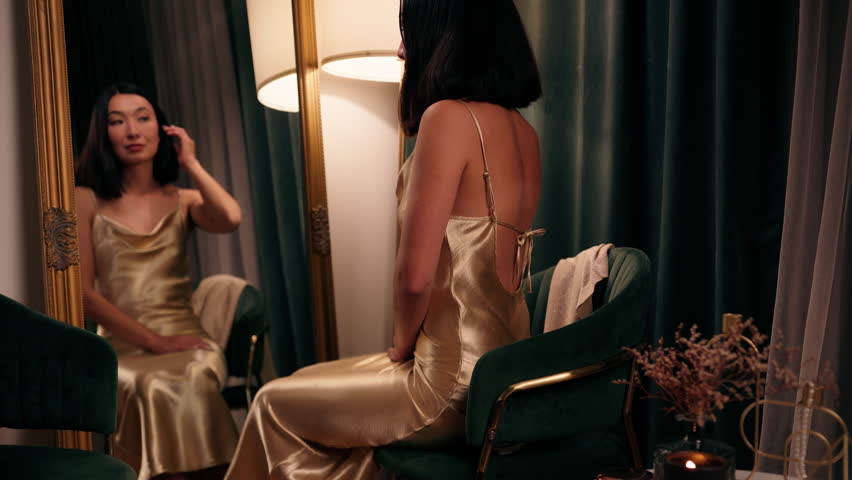 Beautiful elegant Asian woman Japanese Chinese lady looking at mirror reflection touching hair gold stylish dress checking preparing to date party Korean female prepare to event indoors in evening