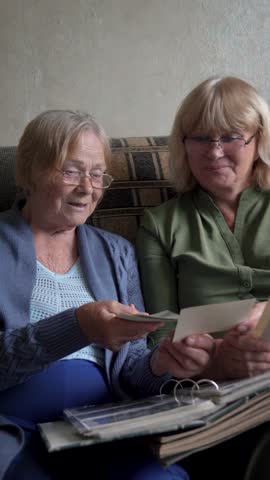 Old female mother with mature woman daughter sitting together on couch in room. Women reminisce while browsing through old retro photos in family album. Relatives gatherings where sharing past stories
