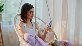 Peaceful brunette reading mobile phone message swinging in cozy rope chair in light apartment interior closeup. Smiling relaxed lady browsing social media at cellphone drinking morning coffee at home. - Powered by Shutterstock - Get 15% off with code: PIKWIZARD15