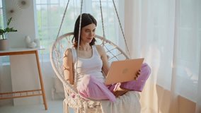 Brunette talking at virtual meeting sitting cross-legged in hanging chair. Smiling woman video calling by laptop relaxing in light apartment. Cheerful lady enjoy friendly online conference in swing. - Powered by Shutterstock - Get 15% off with code: PIKWIZARD15