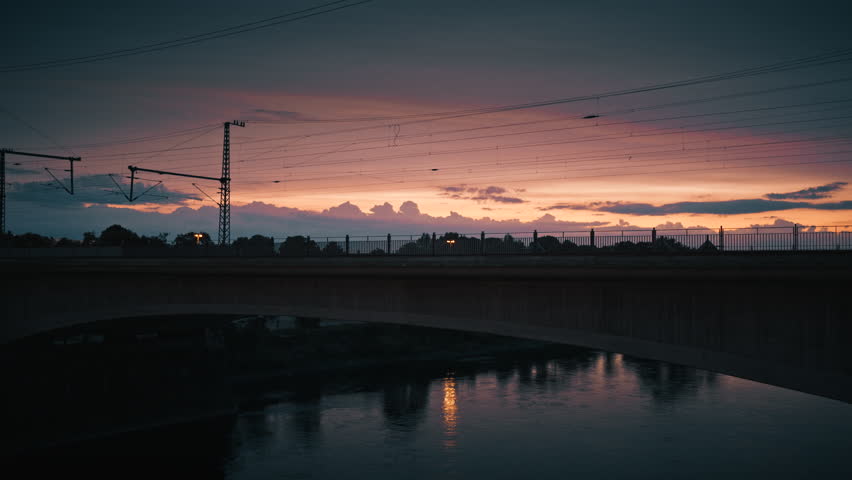 Train crossing a bridge at sunset with dramatic sky colors and its reflection shimmering on the river below.