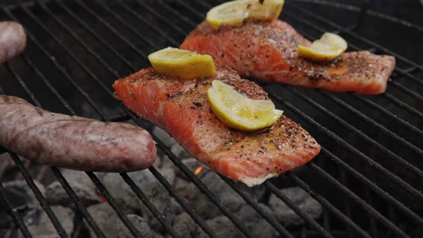 Healthy tasty grilled salmon meat with skin, lemons, olive oil, and spice seasoning grilling on charcoal grill grate with fire and flames at outdoor home cooking of seafood in slo mo and slow motion