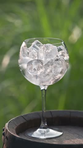 pink sparkling drink pouring in glass with ice from bottle on green summer garden background