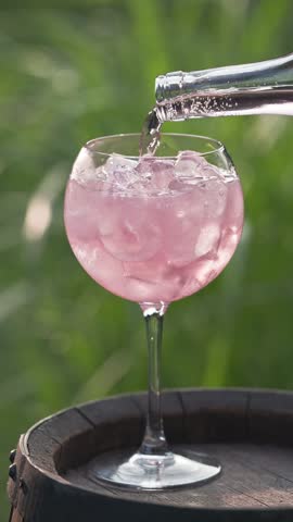 pink sparkling drink pouring in glass with ice from bottle on green summer garden background