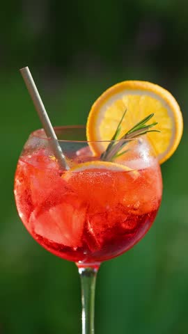 glass of classic red spritz cocktail with ice and orange slices on green garden background