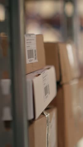 Vertical Video Employee using barcode scanner on cardboard boxes in fulfillment center. Organized shelving, package labels and route planning support distribution and shipment. Logistics hub. Camera B