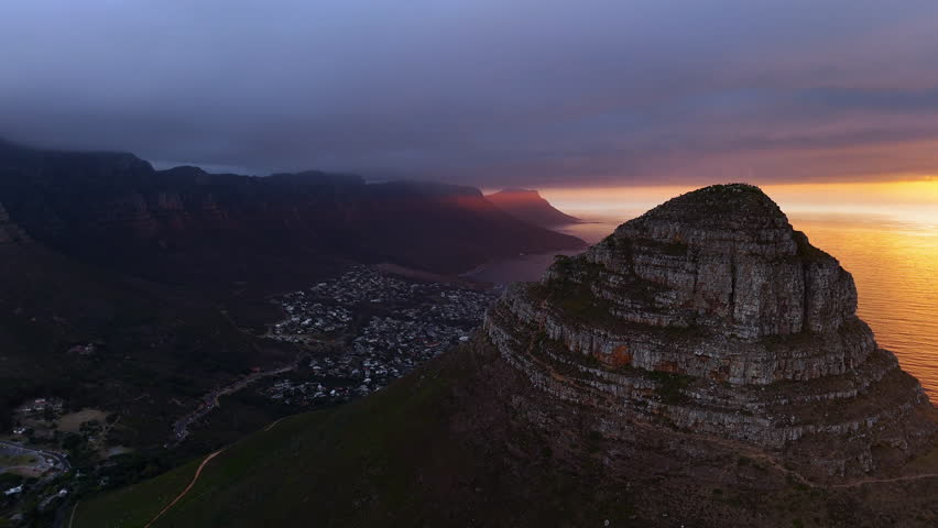 Cinematic aerial view of Table Mountain in Cape Town, South Africa, dramatic rocky cliffs with coastal cityscape at sunset, clouds rolling over peaks and golden evening light