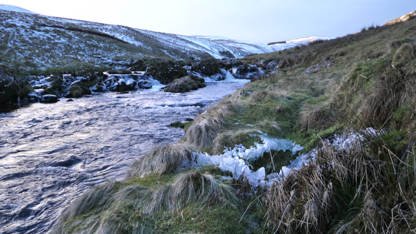 Icy river flowing through snowy hills in Northumberland, UK. Rugged countryside with frozen banks, clear water, and dramatic winter scenery.