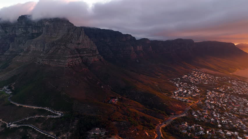 Cinematic aerial view of Table Mountain in Cape Town, South Africa, dramatic rocky cliffs with coastal cityscape at sunset, clouds rolling over peaks and golden evening light