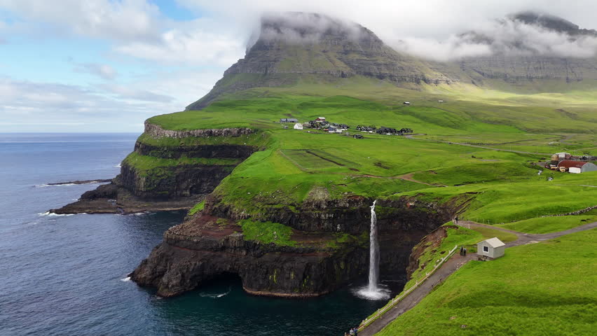 Aerial view of Gásadalur waterfall plunging into the Atlantic Ocean, framed by dramatic cliffs, lush green Faroe Islands landscape, and traditional village houses