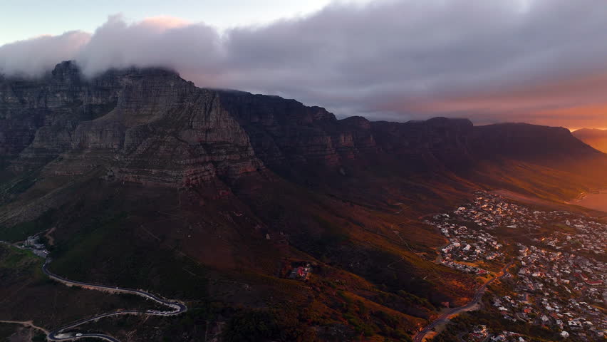 Cinematic aerial view of Table Mountain in Cape Town, South Africa, dramatic rocky cliffs with coastal cityscape at sunset, clouds rolling over peaks and golden evening light