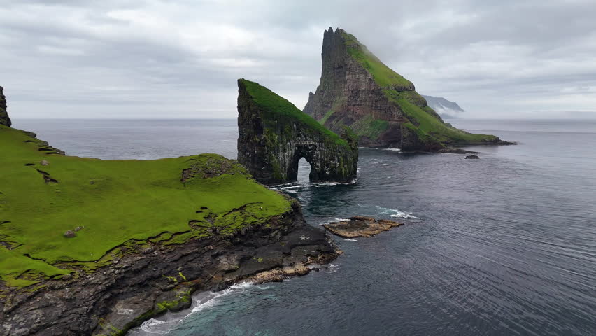 Cinematic aerial view of Drangarnir sea stacks rising dramatically from the Atlantic Ocean near Vágar, Faroe Islands, showcasing rugged cliffs, lush green slopes, and misty Nordic seascape