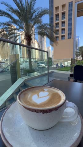 Latte with heart-shaped art served outdoors at Dubai Marina, coffee cup with palm trees, yachts, and skyscrapers in the background, lifestyle and travel vibes.