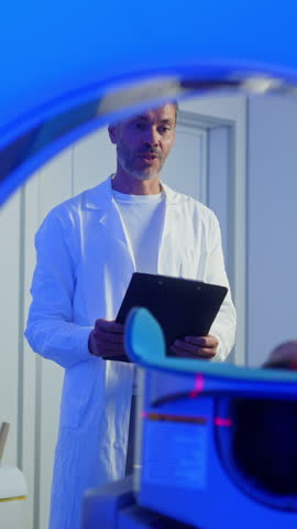 Medical worker standing confidently near MRI tunnel while composed patient lying flat on scanning table. Operating diagnostic system during scheduled medical procedure inside clean examination area.
