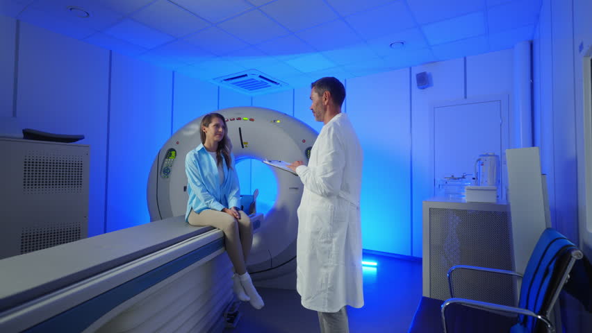 Handsome Caucasian Doctor talking to patient before MRI. Patient talking about symptoms before tomography. Patient sitting at CT scanner doctor adjusting MRI capsule. Conducting advanced imaging