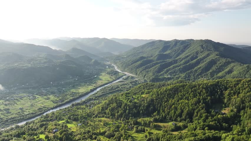 Flight over the summer mountains with mountain river serpentine, green meadows, village and forest. Ukraine, Carpathians