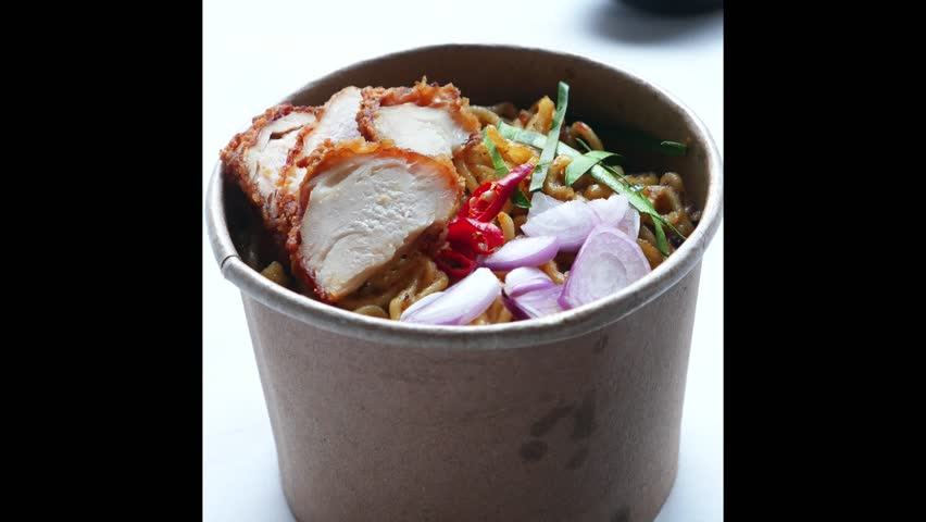 close up, curry noodles topped with fried chicken pieces, served in a paper bowl and chopsticks.